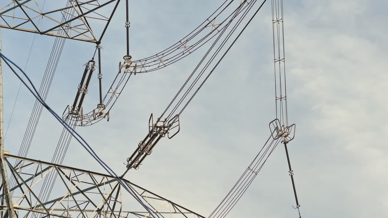 Close-up view of insulators, conductor bundles, and grading or corona rings on a high-voltage transmission tower, steel lattice lines cutting across pale sky in static daylight