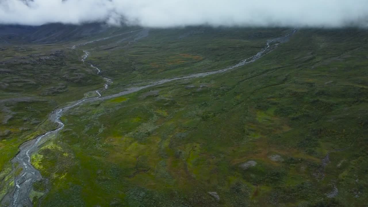 Gorgeous high altitude aerial drone footage view of green colored Sweden terrain landscapes where a small and narrow river flows. Clouds are thick and are low to the ground in the background.