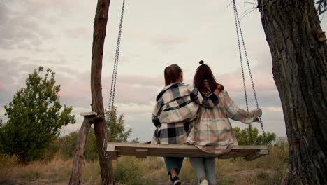 close-up shooting from behind: a brunette girl in a checkered shirt and a blonde girl are swinging on a wooden swing outside the city. rest in the country by the sea