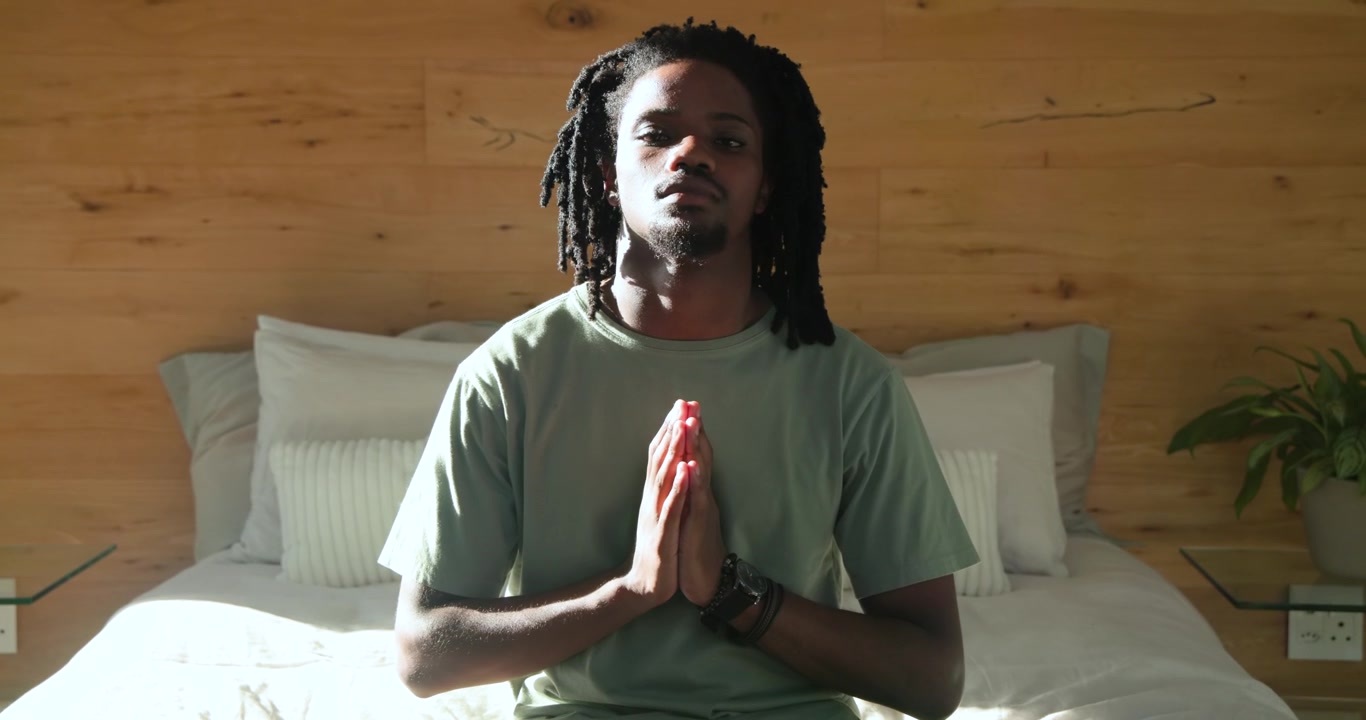 African American man sitting on bed in bedroom praying, ending meditation and looking off-frame