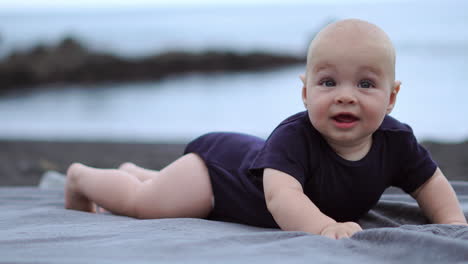 the baby's playful laughter fills the air as he lies on his stomach on the black sand near the ocean, his eyes fixed on the camera