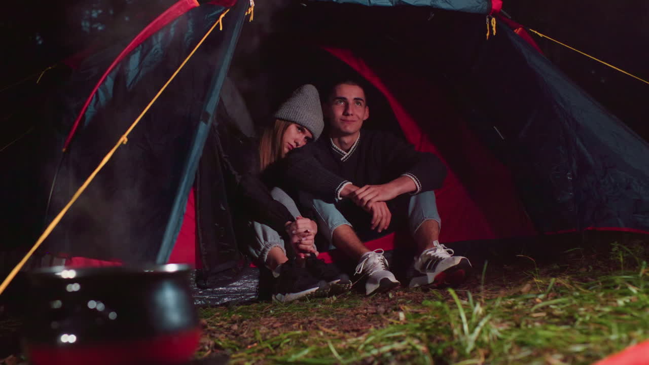 Young couples seated together at entrance of tent during nighttime camping, gazing into distance while steaming pot simmers over fire, surrounded by natural setting and soft glow from warm firelight