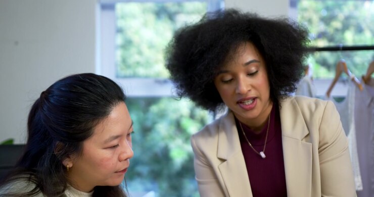 Diverse female team leaning over table seeing samples by clothing rack, discussing designs