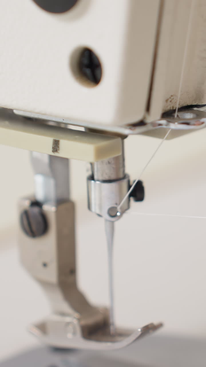 Closeup sewing needle and presser foot on industrial machine, metal components, thin white thread threaded through eye, shallow depth of field, clean studio background, technical precise atmosphere