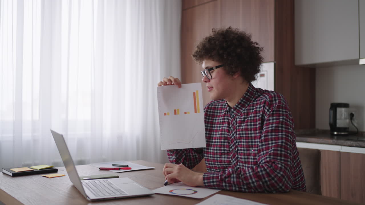 A man with glasses at the table shows a picture with graphs in the camera of the laptop