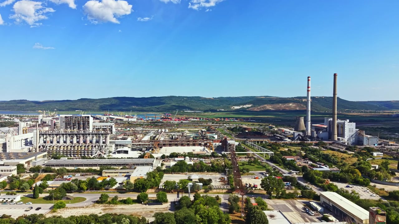 Vista di una fabbrica in una vasta area industriale sotto un cielo azzurro limpido con alberi verdi in primo piano e montagne sullo sfondo.