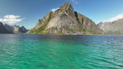 drone flying low over aqua blue water towards a large mountain in lofoten, norway