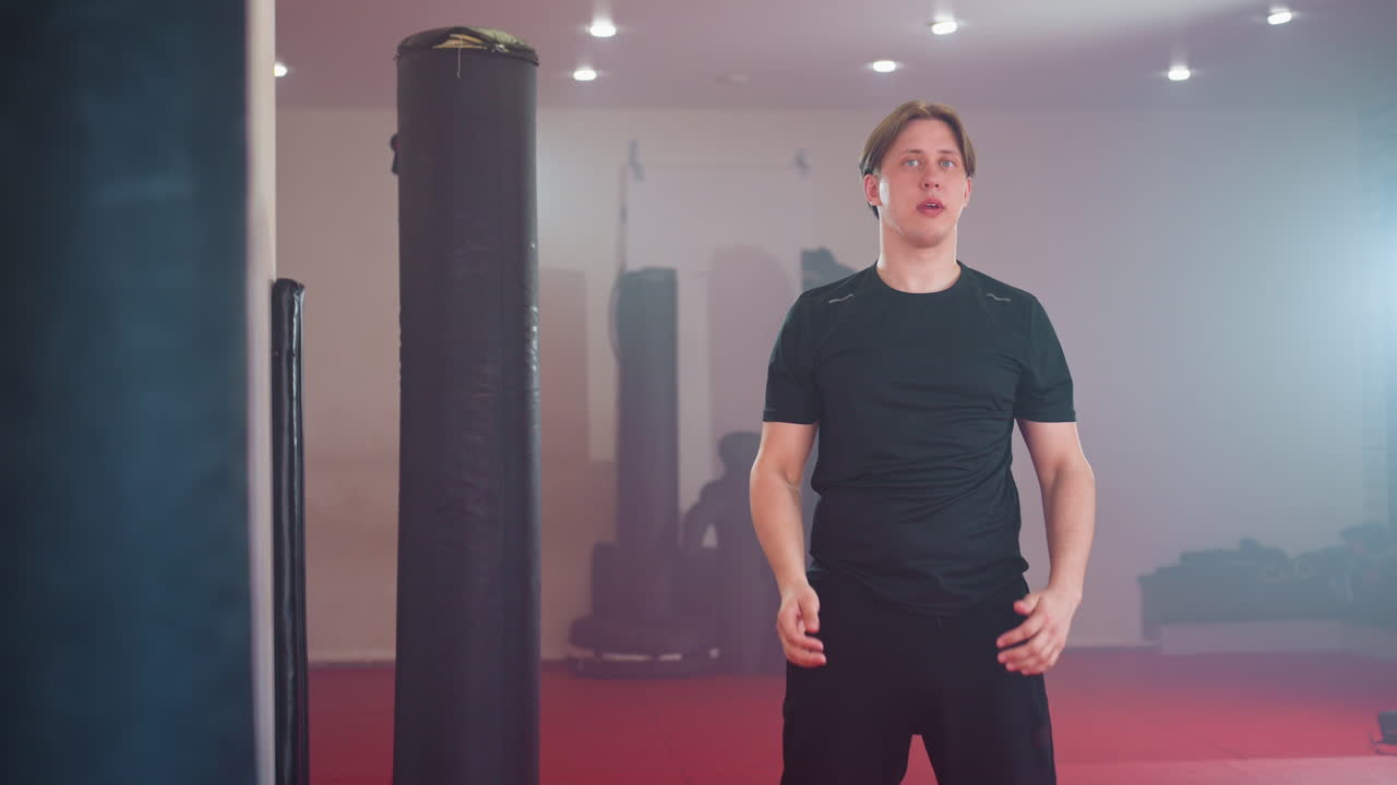 운동하는 남자가 헬스장 환경에서 셔츠를 벗고 강한 조명 아래 샌드백 근처에 서서 운동을 준비하며 가슴과 몸통을 보여주고, 훈련 도전에 대한 강인함과 결의를 나타내요.