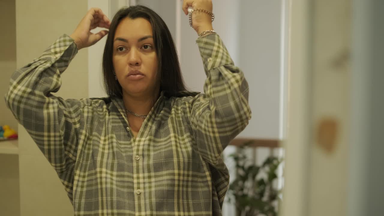 A pregnant woman standing near a mirror at home, putting on clothes. The scene reflects self-care, maternity, and everyday life in a warm and personal setting