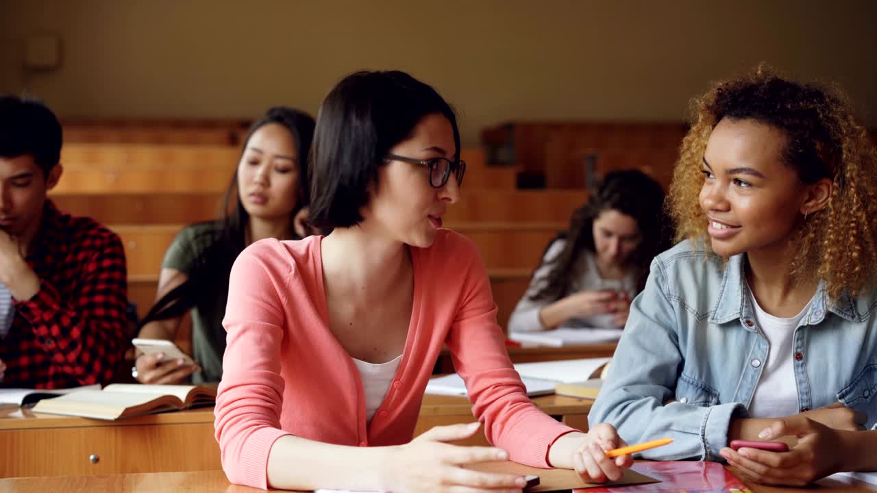 Two friends students are talking and laughing during break between lessons at high school, African American girl is holding smartphone. Communication and people concept.