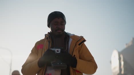 sky view of black man holding camera, looking around on snowy urban street, exhaling visible breath into crisp frosty morning air amid parked cars and tall buildings under pale winter sky