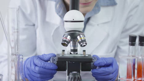 Un-Técnico-De-Laboratorio-Con-Guantes-Azules-Está-Girando-Lentamente-Las-Perillas-De-Un-Microscopio-Con-Tubos-De-Ensayo-Al-Lado