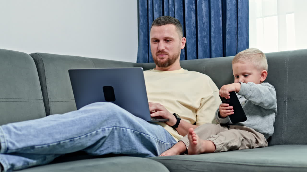 Ojciec i syn siedzą razem na kanapie, korzystając z laptopa i smartfona w jasnym salonie w ciągu dnia.