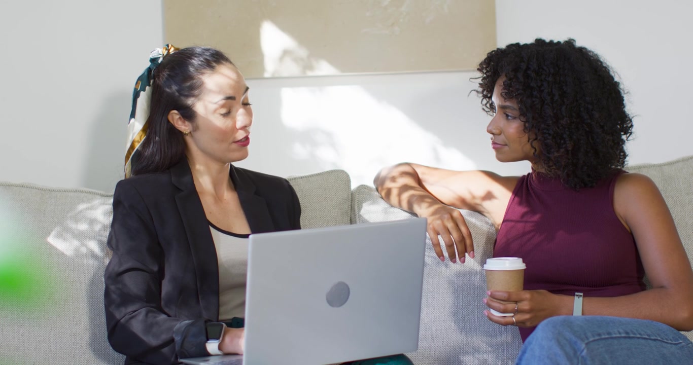 Due donne adulte sedute sul divano, quella a sinistra in blazer posiziona il laptop mentre quella a destra tiene una tazza, mentre rivedono.