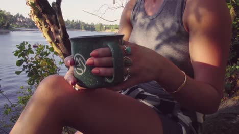 linda chica hipster relajándose con una bebida en una taza de cadera al lado de un río o lago