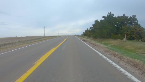 point of view driving on a straight stretch of country highway, past a farm yard and harvested fields