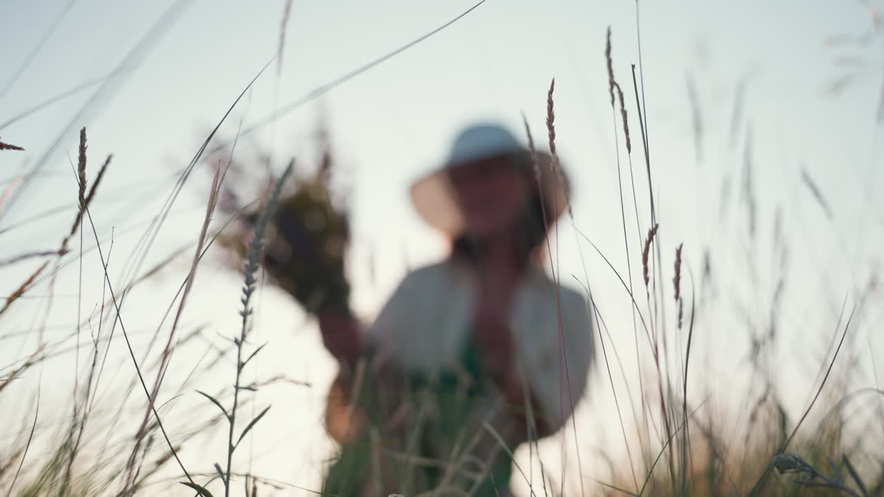 close up of tall wild grass gently swaying in soft breeze with blurred background of woman in hat holding wildflower bouquet, creating dreamy atmosphere filled with calm summer light