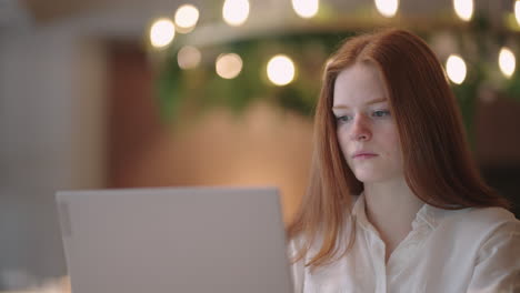 thoughtful brooding remote working red haired woman sitting infront of a laptop or notebook in casual outfit on her work desk in her modern airy bright living room home office with many windows