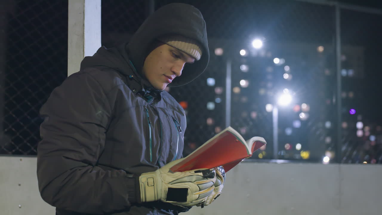 후디와 베니를 입은 골키퍼가 메탈 골포스트에 기대어서 도시 불빛 아래 야외에서 책을 사려 깊게 읽는 클로즈업
