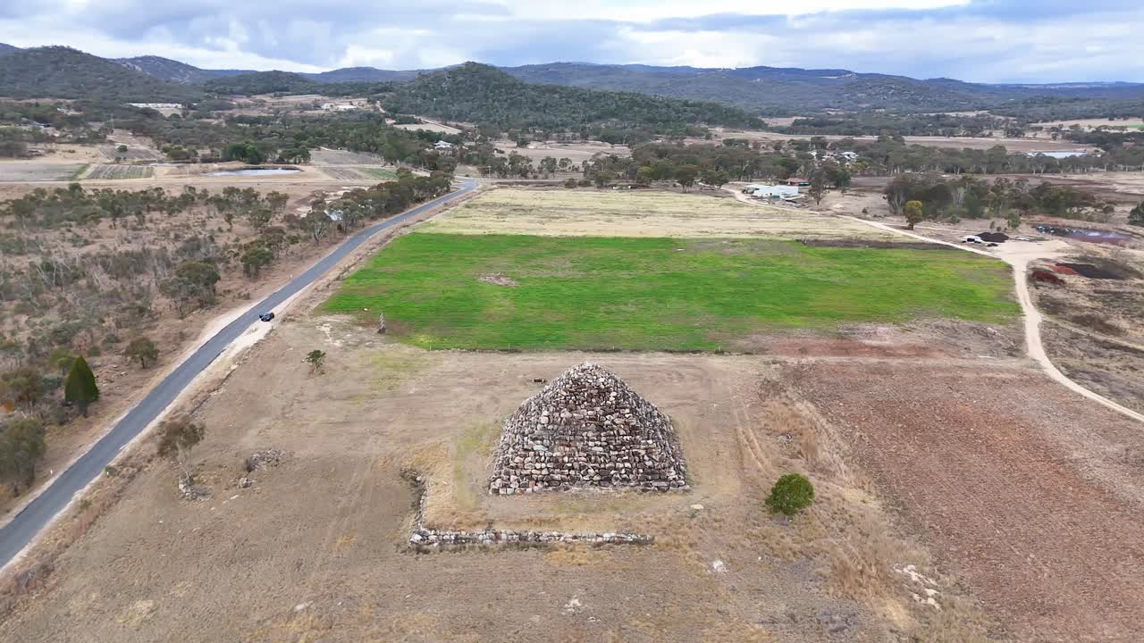 Drone footage circles a large granite rock pyramid in a dry, rural landscape near Ballandean, Australia. Overcast daylight, smooth camera movement