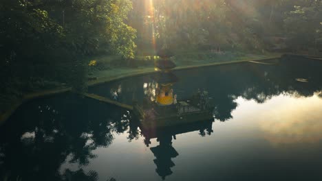 Woman-walking-towards-Bali-temple-in-pond-with-magical-morning-sunlight,-aerial