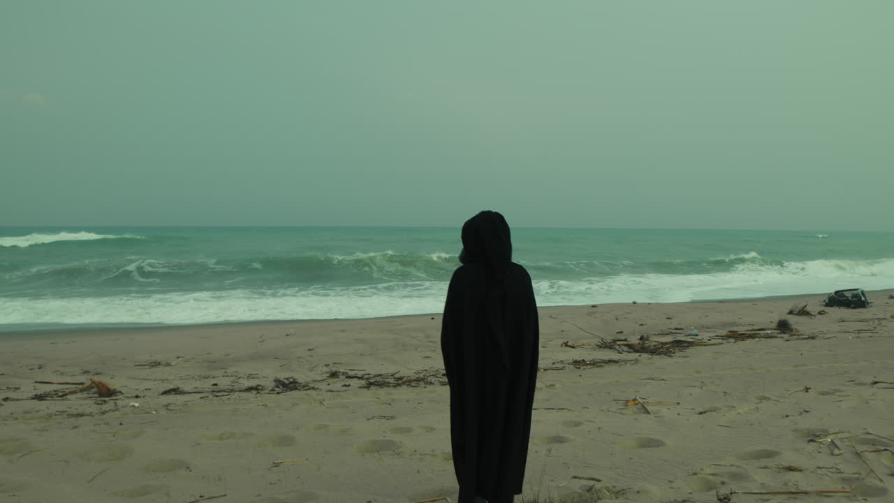 Man Walks Towards Stormy Ocean Wearing Black Cloak Over Shoulders