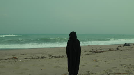 man walks towards stormy ocean wearing black cloak over shoulders