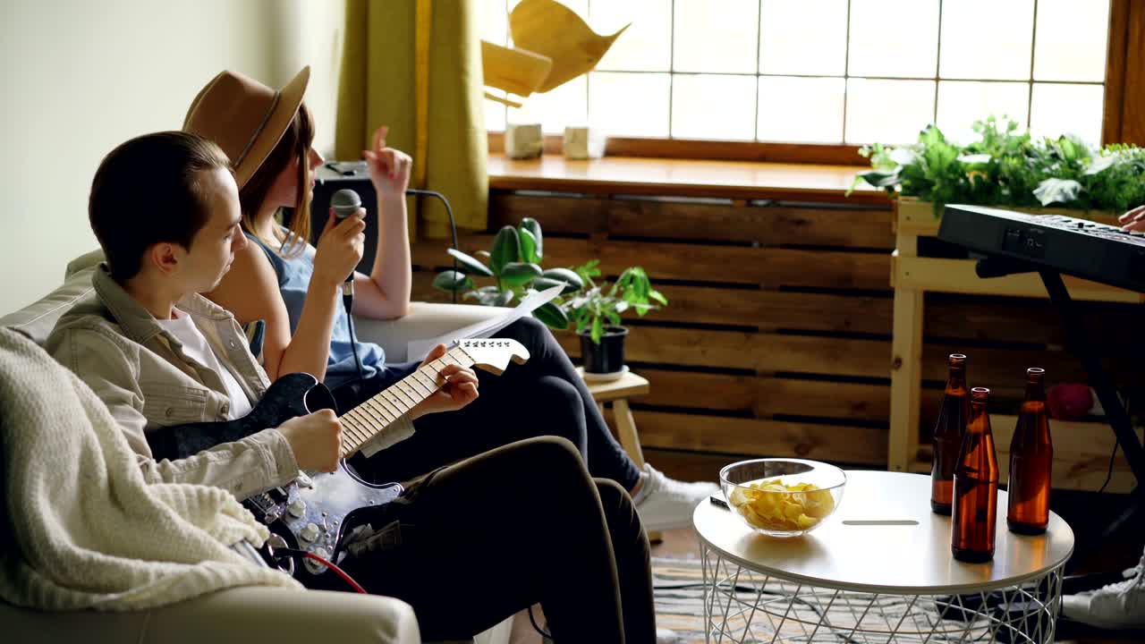 쾌활한 젊은 음악 밴드가 음악 악기 기타와 키보드를 연주하고 좋은 집에서 리허설 중에 마이크에서 노래하고 있습니다.