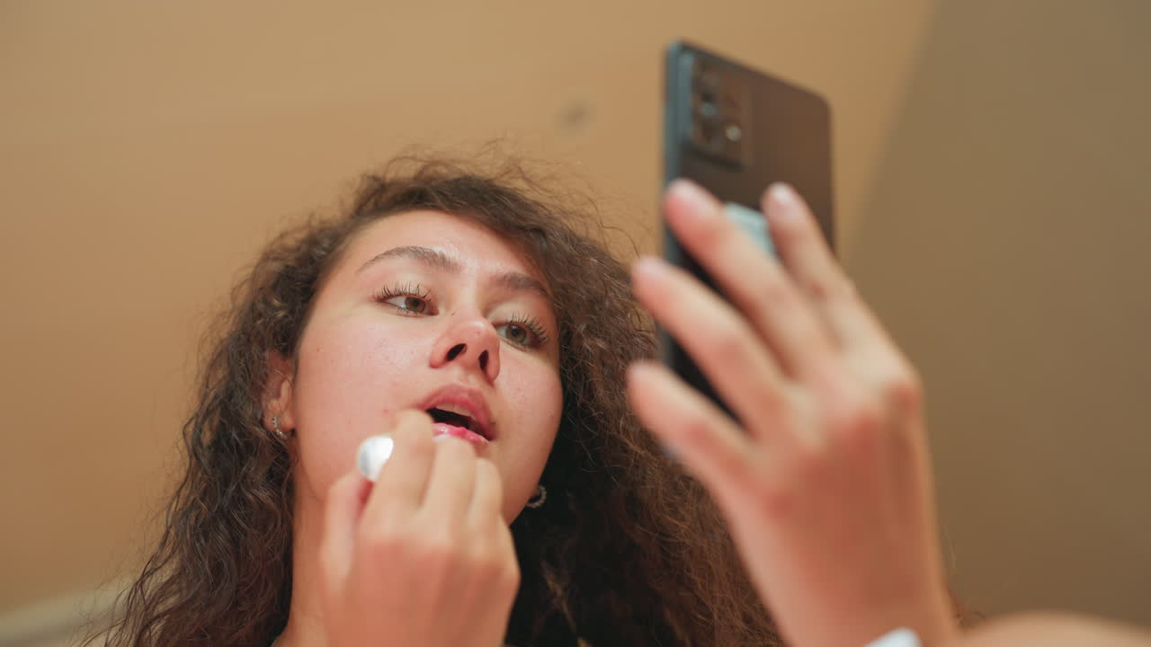 Influencer applying gloss. Latina creator using lip balm. Close view of influencer applying lip gloss beside mirror. Woman in hotel room applying lip product while examining her reflection