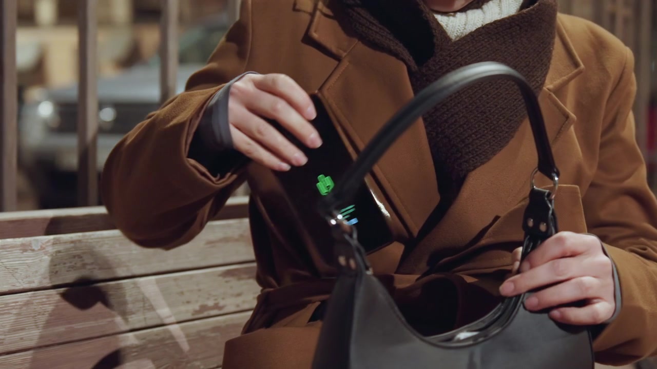 Donna su una panchina che trova il telefono nella borsa, momento di sollievo in un parco serale con cinturino in pelle e bagliore dello schermo, primo piano delle dita che estraggono il dispositivo dalla tasca, cappotto marrone accogliente e atmosfera di luce del lampione.
