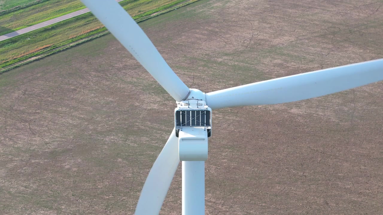 Primo piano di una turbina eolica che gira nel vento, con campi marroni e verdi intorno, di giorno, vista posteriore, ripresa dal basso
