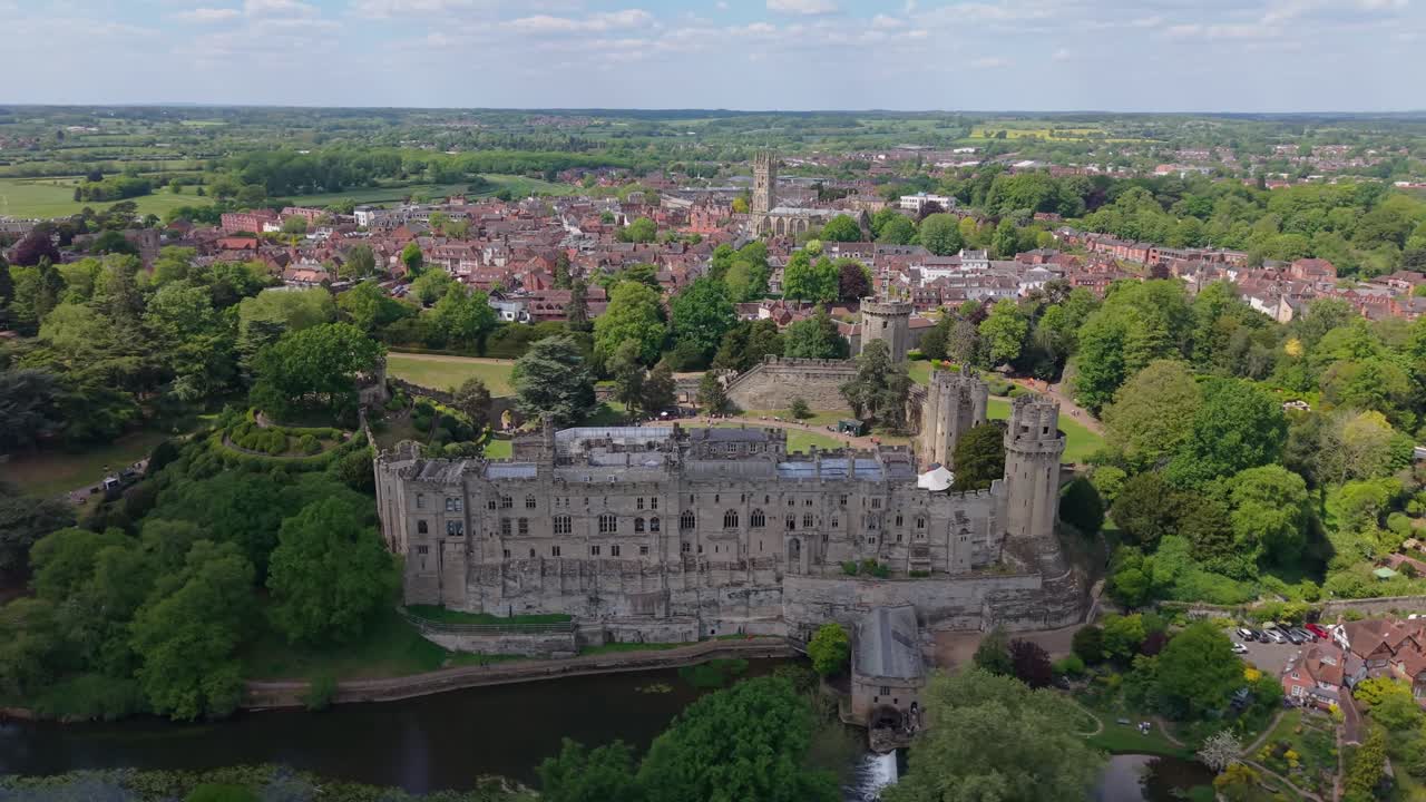 Un drone orbita sopra il Castello di Warwick a Warkside, rivelando la fortezza storica con il pittoresco villaggio e il paesaggio campestre che si estendono sullo sfondo.