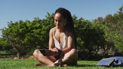 mujer afroamericana sentada en la hierba usando un teléfono inteligente y mirando un reloj inteligente en el parque