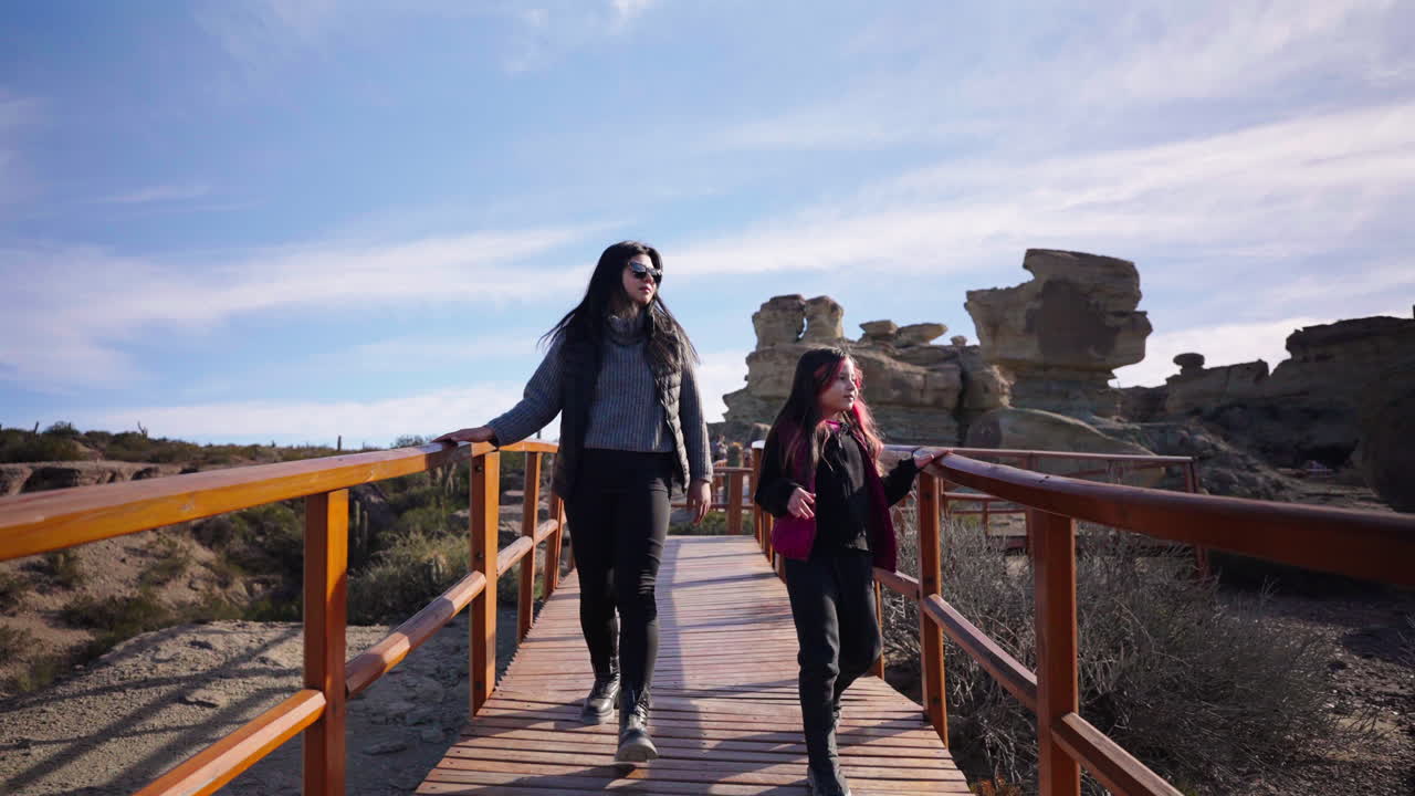 Small family walking along wooden path surrounded by rocky formations under bright sky at Ischigualasto Park Valley of the Moon, Argentina