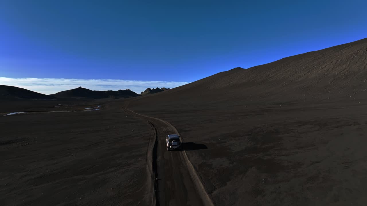Inquadratura aerea con drone di un fuoristrada che percorre una strada sterrata solitaria attraverso il deserto vulcanico nero dell'Islanda sotto un cielo blu intenso. Viaggio epico e remoto, solitudine e avventura fuori dai sentieri battuti