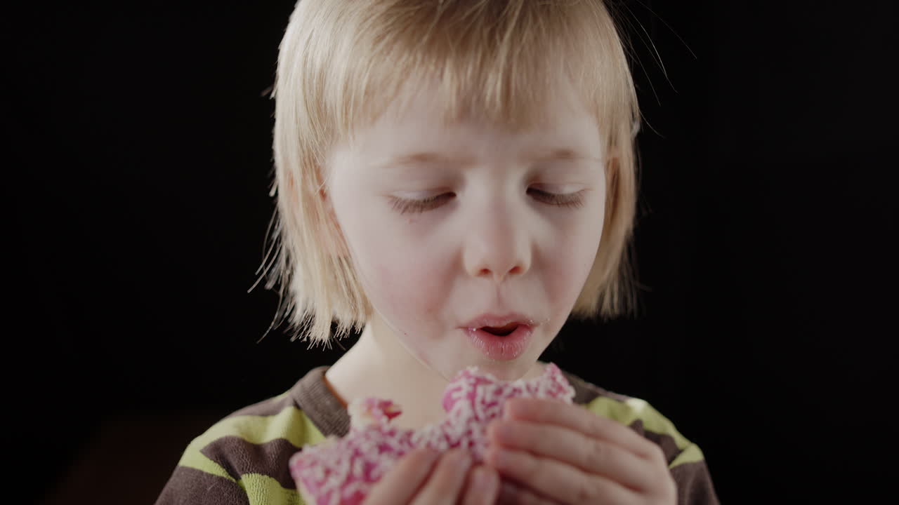 Cute caucasian girl bites into yummy pink glazed donut with content expression
