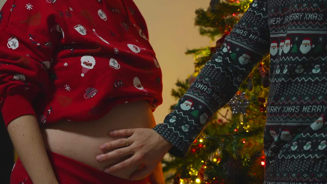 Foto de close-up de um casal grávido vestindo roupas natalinas, tentando sentir os chutes do bebê em frente à árvore de Natal iluminada