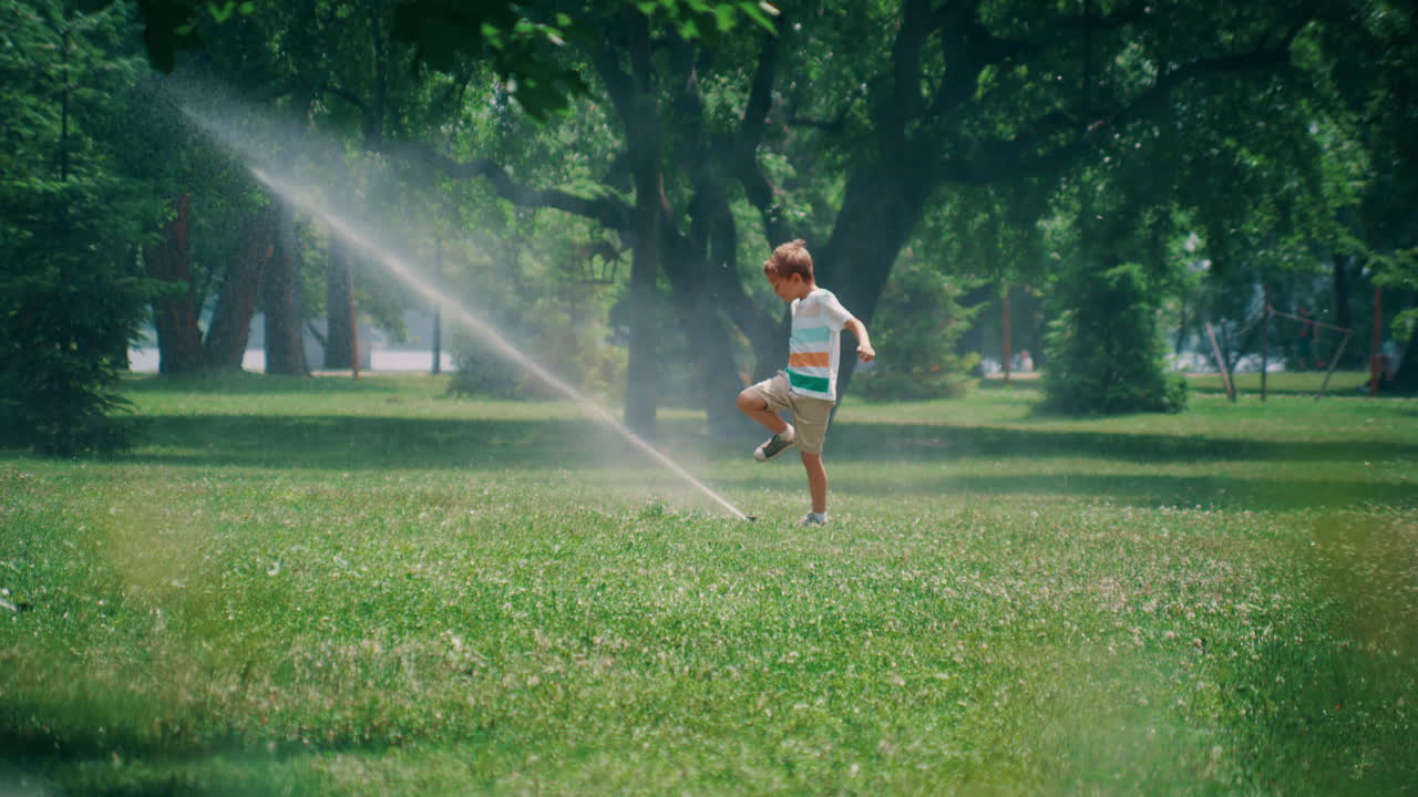 Playful elementary age boy kicking water sprinkler jet. Kid enjoy time in park