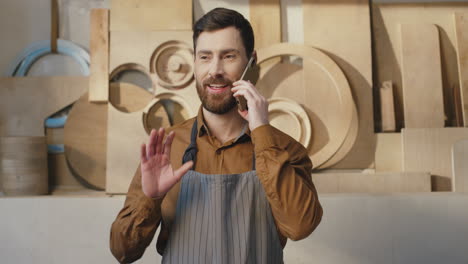 portrait of caucasian bearded man in apron talking on the phone in carpentry workshop