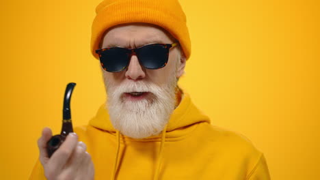 Confident-aged-man-smoking-pipe-indoors.-Stylish-old-guy-speaking-on-camera.