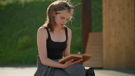 lady seated with legs crossed reading a book while the wind blows her hair, a black bag sits beside her, and the blurred background features greenery and wooden structures