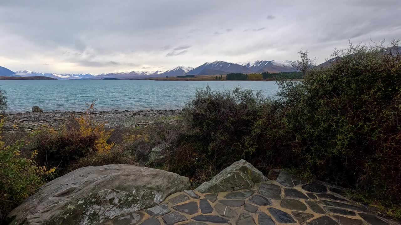 Camera slowly pans across stone terrace to reveal Lake Tekapo, rocky shoreline, and distant snow-capped mountains under overcast daylight