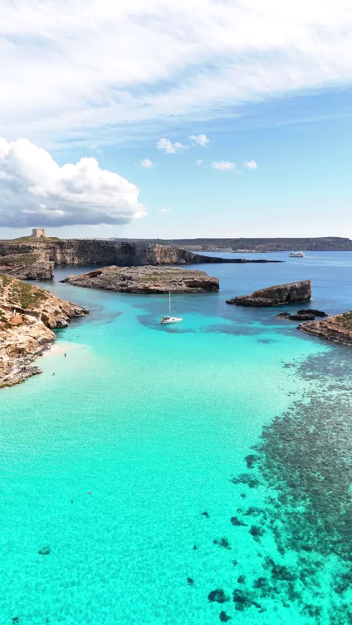 Veduta aerea della Blue Lagoon a Comino, Malta, con diverse barche ancorate su acqua turchese vicino a una spiaggia sabbiosa e una costa rocciosa sotto nuvole sparse