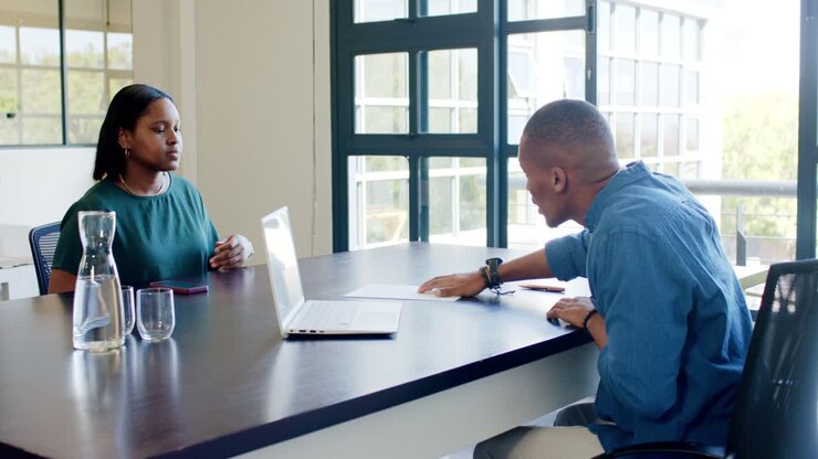 African American coworkers in meeting man blue shirt sliding paper, woman green top reading laptop