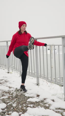 woman holding iron railing stretching leg back and forth on snowy winter ground amid serene foggy landscape with bare tree silhouettes and railing overlooking misty horizon