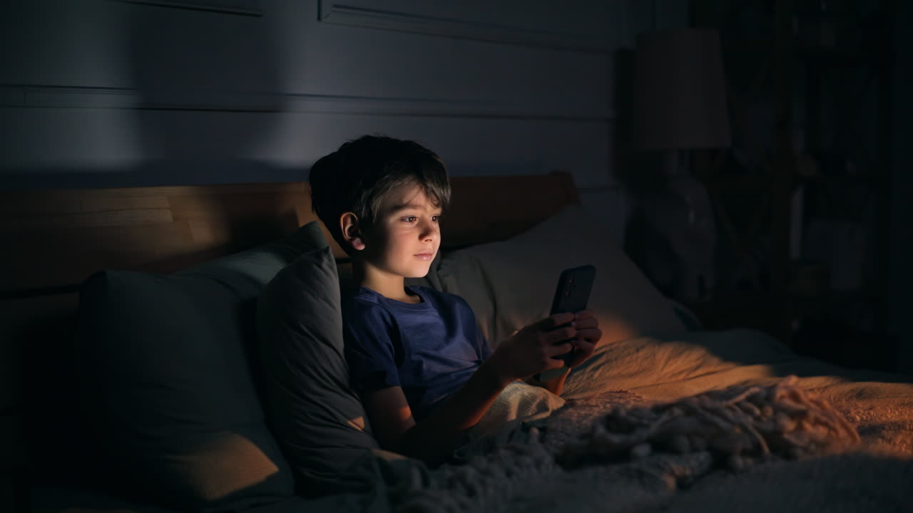 Little kid looking cellphone lying dark bedroom closeup. Boy staring smartphone