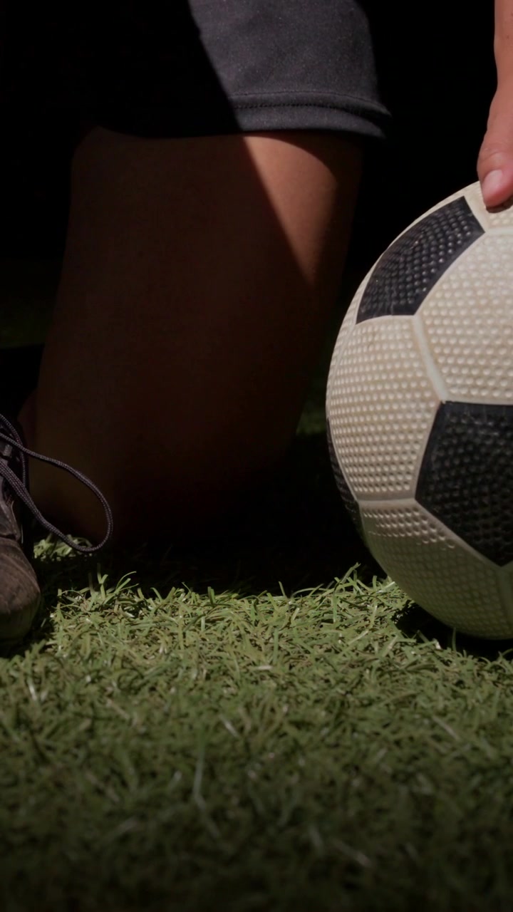 Video vertical: Jugador arrodillado con tacos, atándose los cordones en césped artificial, estabilizando el balón para jugar.