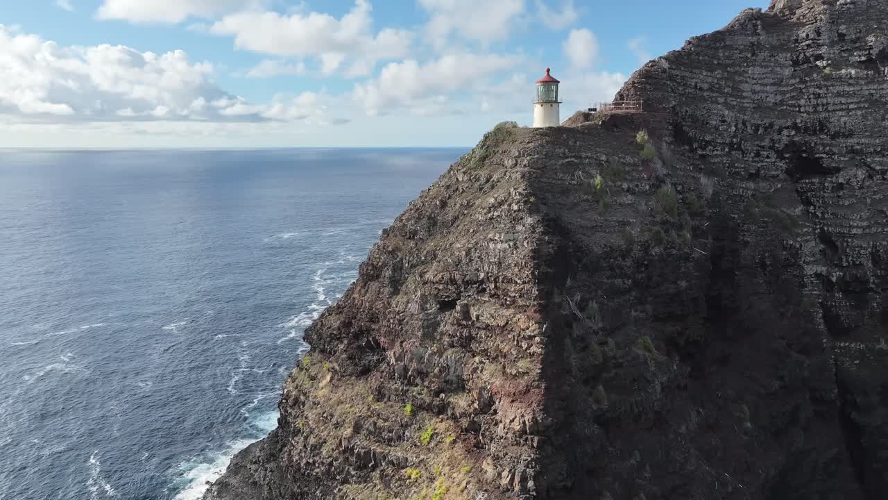 Aerial drone footage of a lighthouse perched on Oahu’s windward coast cliffs, Hawaii, showcasing dramatic rocky coastline, turquoise ocean waves, lush tropical vegetation, and panoramic ocean views