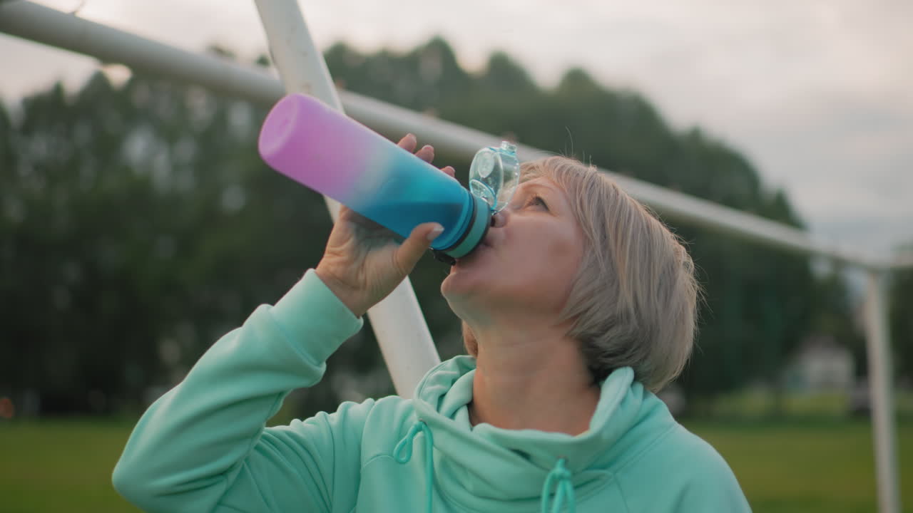 Female athlete in blue hoodie drinks water from vibrant colorful bottle, standing relaxed by goalpost in field during peaceful morning workout, enjoying hydration with refreshing satisfaction