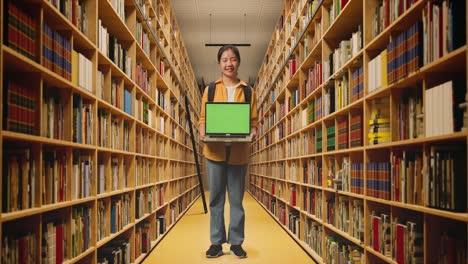 cuerpo lleno de estudiante mujer asiática con una mochila sonriendo y mostrando pantalla verde portátil a la cámara mientras estaba de pie en la biblioteca